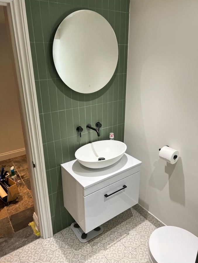Olive tiled wall with round mirror, white countertop basin and black wall tap