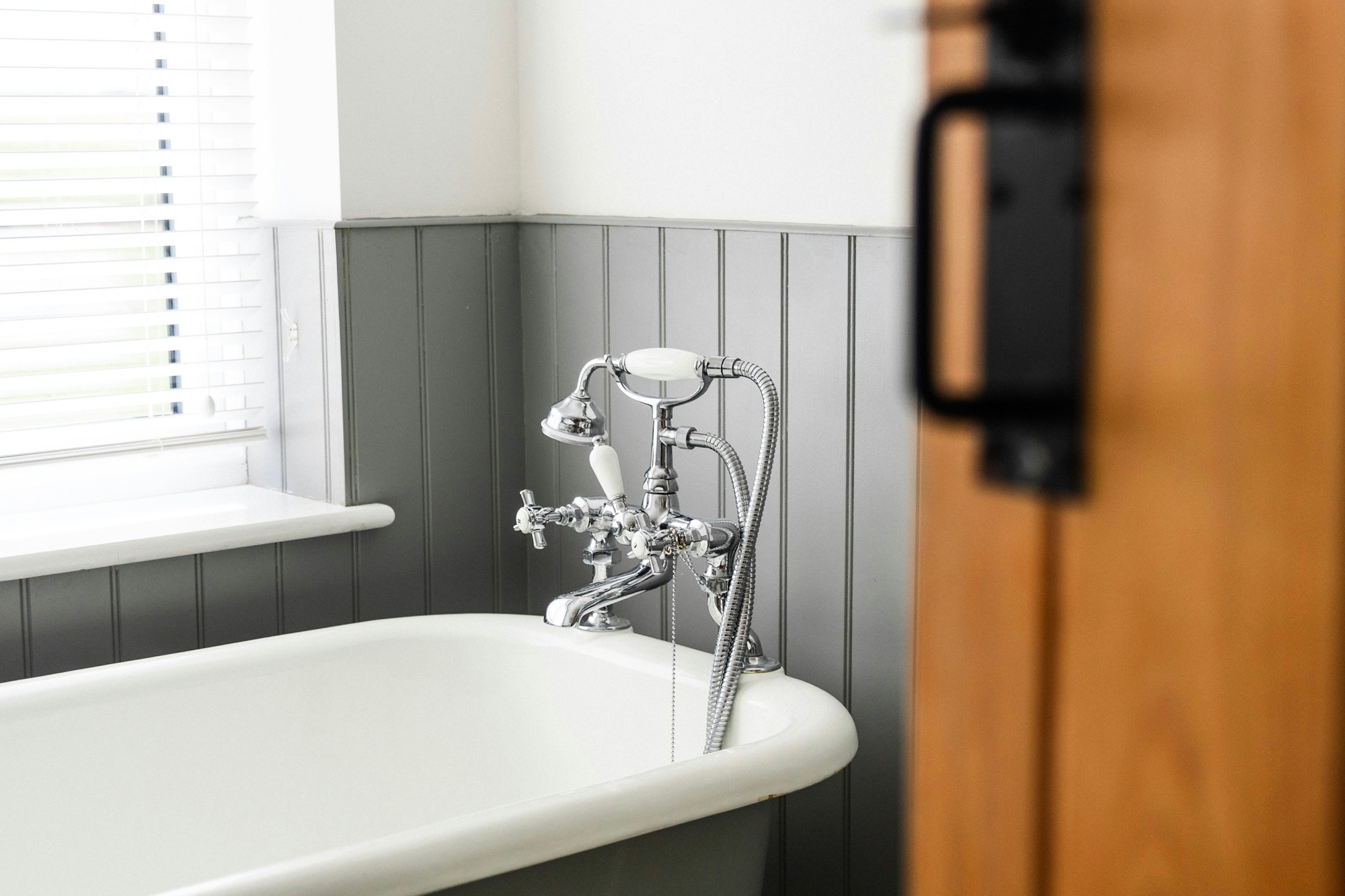 A roll-top bath against grey panelling with a brass shower mixer, lit by a sash window